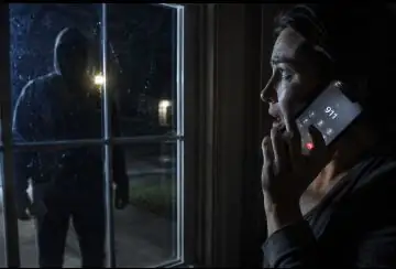 A dark tense nighttime scene showing a shadowy figure outside a house window with the homeowner inside looking alarmed and calling emergency services on a mobile phone, dramatic shadows and light from phone screen, photorealistic, cinematic.