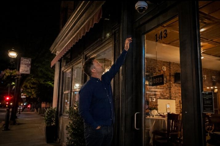 A restaurant owner in his 40s standing outside his closed restaurant at night on a quiet US city street, checking a mounted security camera above the entrance.